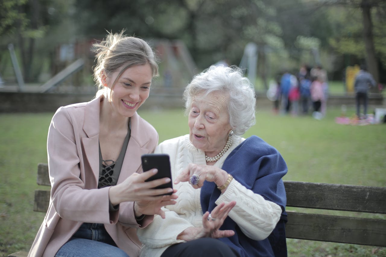 service-01 Delighted female relatives sitting together on wooden bench in park and browsing mobile phone while learning using